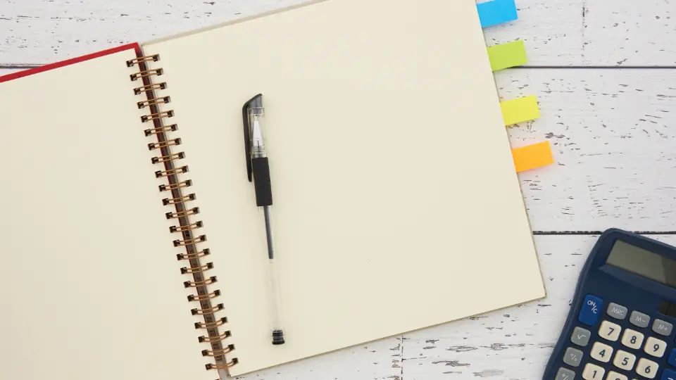Beyaz ahşap masada açık spiral defter, tükenmez kalem ve hesap makinesi